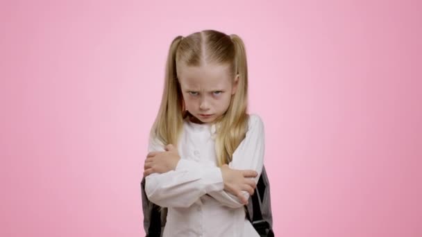 Victim of bullying. Portrait of little grumpy schoolgirl with backpack posing with crossed arms and looking sullenly to camera, feeling offended, pink studio background, slow motion - Footage, Video