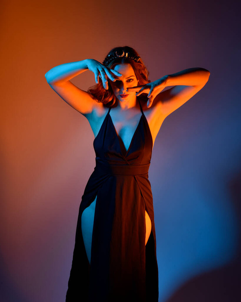 Κοντινό πορτρέτο του όμορφου γυναικείου μοντέλου που φοράει κομψό μαύρο φόρεμα, ποζάρει σε φόντο στούντιο με χειρονομίες εμπνευσμένες από τη φαντασία, πολύχρωμο δημιουργικό φωτισμό. - Φωτογραφία, εικόνα