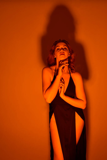 Κοντινό πορτρέτο της όμορφης γυναίκας μοντέλο φορώντας κομψό μαύρο φόρεμα και στέμμα, ποζάροντας σε φόντο στούντιο με φαντασία εμπνευσμένη χειρονομίες, πολύχρωμο χρυσό δημιουργικό φωτισμό. - Φωτογραφία, εικόνα
