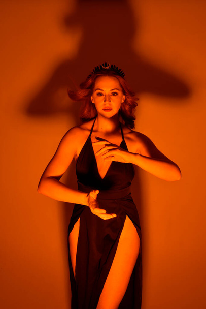 Portrait en gros plan d'une belle mannequin femme portant une élégante robe noire et une couronne, posant sur un fond de studio avec des gestes de bras inspirés de fantaisie, un éclairage créatif doré multicolore. - Photo, image