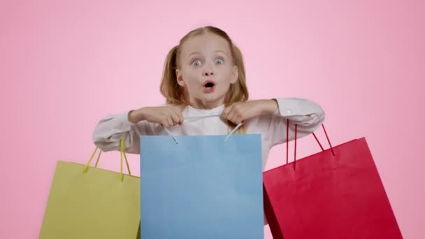 Ωραία ψώνια για παιδιά. Studio πορτρέτο του συγκλονισμένοι ενθουσιασμένοι κοριτσάκι shopaholic απολαμβάνοντας αγορές με πολύχρωμες τσάντες shopper και γέλιο, ροζ φόντο, αργή κίνηση - Πλάνα, βίντεο