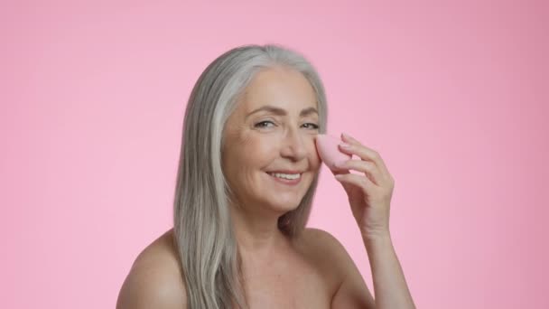 Secretos de belleza. Retrato de estudio de mujer mayor de pelo gris feliz aplicando corrector con esponja de maquillaje especial, sonriendo a la cámara, fondo rosa, cámara lenta - Imágenes, Vídeo