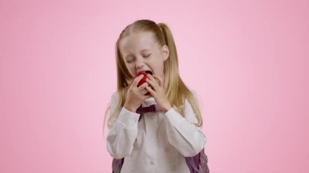 Snack orgánico saludable. Pequeña colegiala feliz con mochila mordiendo manzana fresca madura, disfrutando de fruta, fondo de estudio rosa, cámara lenta - Metraje, vídeo