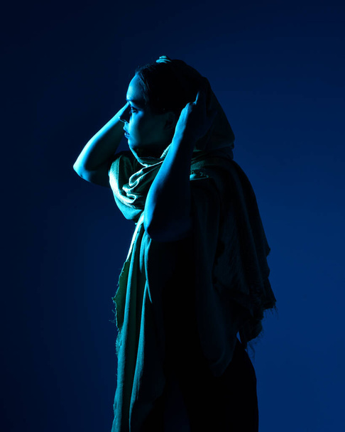 Κοντινό πορτρέτο της όμορφης γυναίκας μοντέλο φορώντας κομψό μαύρο φόρεμα, κουκούλα μανδύα, ποζάρουν σε φόντο στούντιο με φαντασία εμπνευσμένη χειρονομίες, πολύχρωμο δημιουργικό φωτισμό. - Φωτογραφία, εικόνα