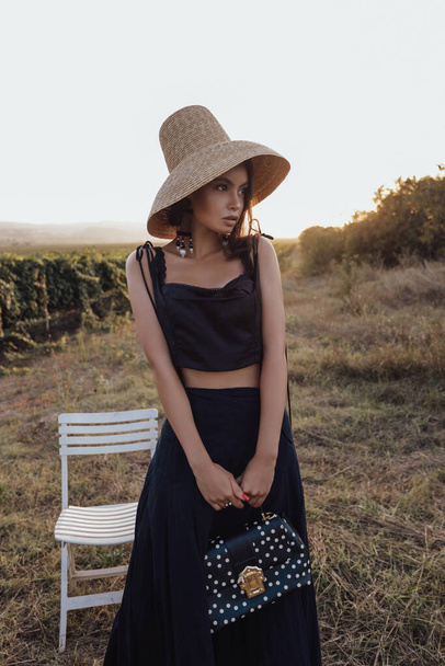 fashion outdoor photo of beautiful woman with dark hair in elegant dress and straw hat posing in the vineyard on the sunset - Photo, Image