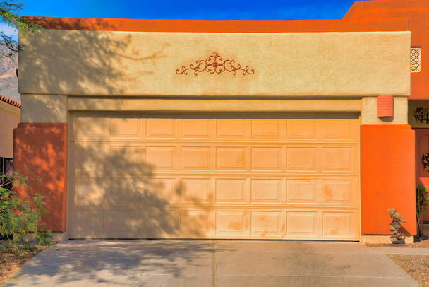 Большая секционная дверь гаража с бетонным подъездом в Тусоне, Аризона. Внешний вид гаража с желтой и оранжевой стеной рядом с входной дверью справа. - Фото, изображение