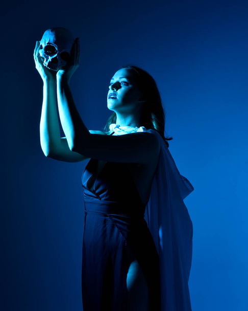 Κοντινό πορτρέτο της όμορφης γυναίκας μοντέλο φορώντας κομψό μαύρο φόρεμα και κουκούλα μανδύα, κρατώντας ένα κρανίο. ποζάροντας σε φόντο στούντιο με φαντασία εμπνευσμένες χειρονομίες, πολύχρωμο δημιουργικό φωτισμό. - Φωτογραφία, εικόνα