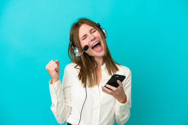 Telemarketer Anglická žena pracující se sluchátky izolované na modrém pozadí s telefonem ve vítězné pozici - Fotografie, Obrázek