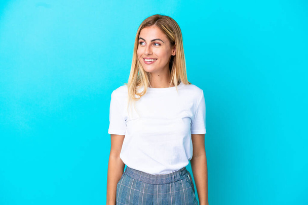 Блондинка Уругвая девушка изолированы на синем фоне, глядя в сторону и улыбаясь - Фото, изображение