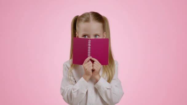 Educazione, ritorno al concetto di scuola. Ragazzina giocosa che copre il viso con notebook e sbirciare fuori, guardando la fotocamera, sfondo rosa studio, rallentatore - Filmati, video