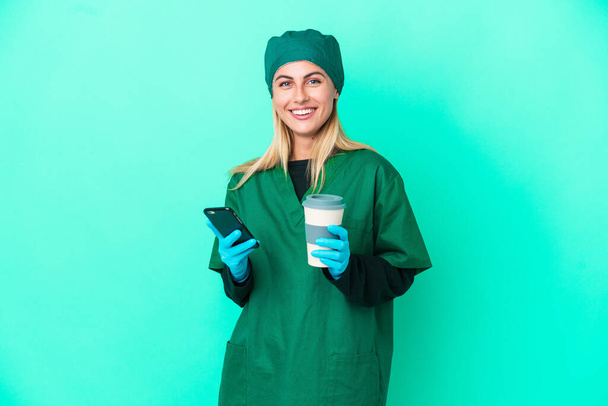 Jonge chirurg Uruguayaanse vrouw in groen uniform geïsoleerd op blauwe achtergrond met koffie om mee te nemen en een mobiele - Foto, afbeelding