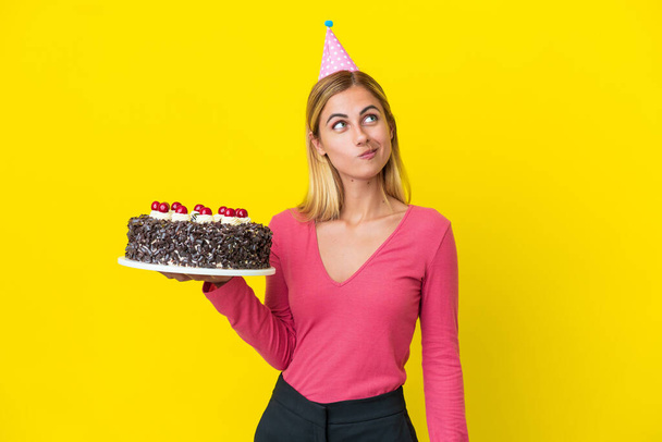 Blonde Uruguayan girl holding birthday cake isolated on yellow background and looking up - Foto, imagen