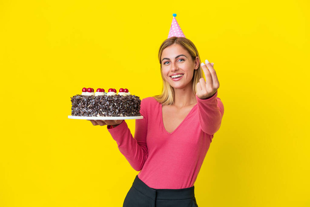 Блондинка Уругвая держит торт на день рождения изолирован на желтом фоне, зарабатывая деньги жест - Фото, изображение