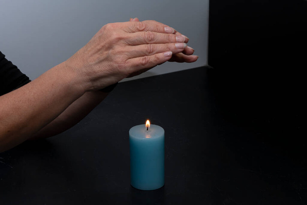 Vanhempi nainen lämmittää kätensä yli kynttilän liekki, valikoiva keskittyä. Käsite: lämmitys- ja sähkökatkokset köyhyysrajan alapuolella. - Valokuva, kuva
