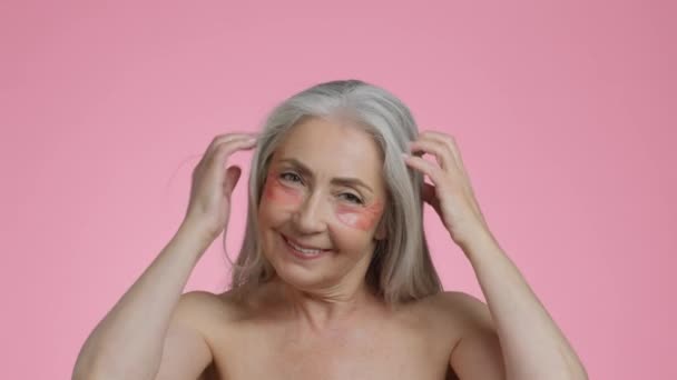 Ochtendschoonheidsrituelen. Studio portret van zorgeloze senior dame met anti-oedeem hydrohel pleisters onder de ogen spelen met haar grijze haar, glimlachen voor de camera, roze bakgrond, slow motion - Video