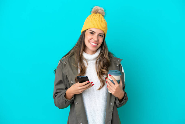 Jovem italiana vestindo casaco de inverno e chapéu isolado no fundo azul segurando café para levar e um celular - Foto, Imagem