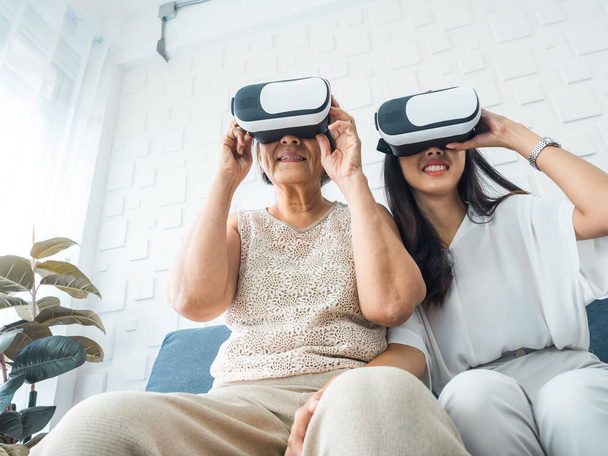Asian old senior woman, mother and happy young daughter, female wearing VR glasses, enjoy 3d game online together in white room. Mom and daughter with reality virtual technology lifestyle concept. - Photo, Image