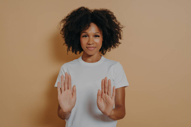 Slow down. Young smiling mixed race woman with curly hairstyle raising palms in stop or prohibition gesture and saying no while standing dressed in white t-shirt over brown studio wall - Photo, Image