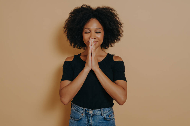 Afro American young woman praying and holding hands in prayer gesture standing isolated on dark beige studio background with copy space, smiling with eyes closed, Praying and hope concept - Foto, Imagen