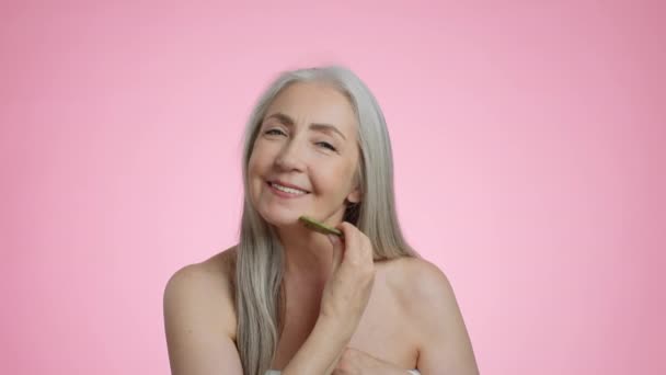 Öregedés elleni szépségápolás. Közelkép portré meglehetősen pozitív ősz hajú idős hölgy csinál önálló masszázs gouache kő, élvezi a bőr emelés eljárás, rózsaszín stúdió háttér, lassított felvétel - Felvétel, videó