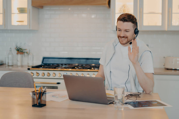 Positive male freelancer using laptop at home during online conference with colleagues while waving to them and looking at screen, sitting at his workplace at home office. Freelance concept - Φωτογραφία, εικόνα