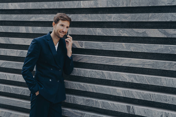 Unshaven male corporate worker dressed in formal black suit keeps hand in pocket has telephone conversation stands near marble wall outdoor. Handsome CEO communicates with professional partner - Fotografie, Obrázek