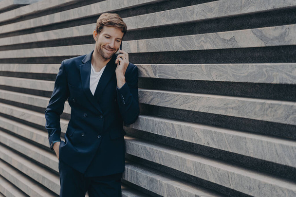 Half length shot of successful businessman calls via smartphone during work break talks with business partner dressed in formal clothes stands near marble wall copy space for your promotion. - Фото, изображение