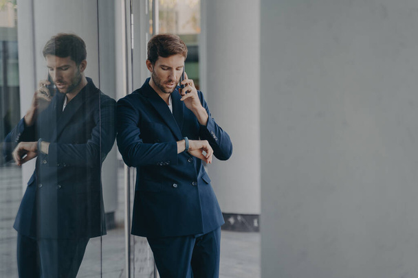 Pensive young businessman in formal suit talking on mobile phone standing outside of office center entrance while hastily glancing at his hand watch, worried about being on time for company meeting - 写真・画像