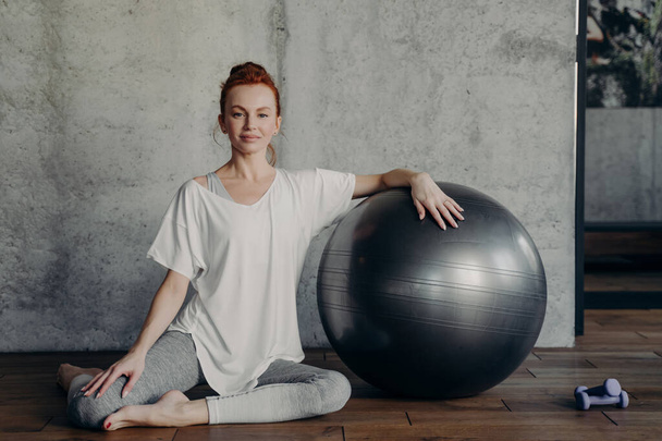 Portrait of beautiful sportive red haired lady gracefully sits on wooden floor in fitness or pilates studio and smiling happily at camera while leaning with one hand on fitball next to dumbbells - Foto, Imagem