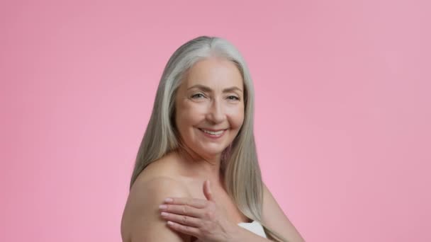 Φροντίδα σώματος και γήρανση. Όμορφη γκρίζα μαλλιά ηλικιωμένη κυρία εφαρμογή κρέμα περιποίησης στο γυμνό ώμο της, τρίψιμο καλλυντικά, ροζ φόντο στούντιο, αργή κίνηση, ελεύθερο χώρο - Πλάνα, βίντεο