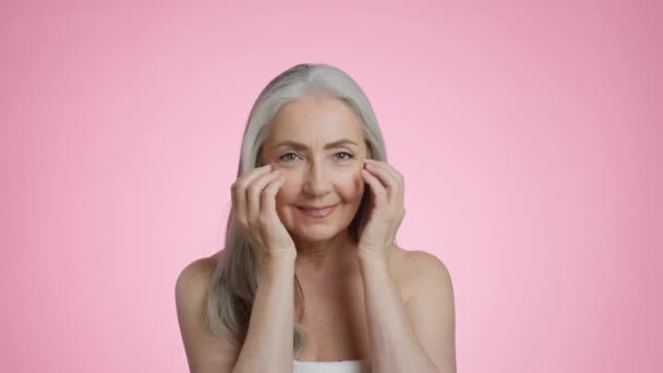 Natuurlijke oude schoonheid. Studio portret van vrij senior grijs harige dame gewikkeld in handdoek aanraken van haar gezicht, glimlachen om de camera, roze achtergrond, slow motion - Video