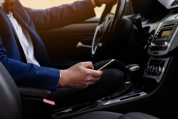 Obchodník v obleku sedí na sedadle řidiče svého luxusního auta a píše sms na smartphone. Mladý muž bere zastavit pro kontrolu pracovních e-mailů na mobilní telefon v pohodlném autě při jízdě do kanceláře. - Fotografie, Obrázek
