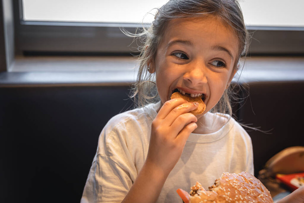 Маленька дівчинка в кафе їсть фастфуд, бургер і картоплю фрі
. - Фото, зображення
