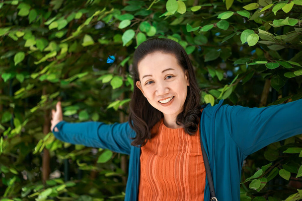 Frau breitet die Arme aus und lächelt mit Kamera vor grünen Büschen. Sie trug orangefarbenes Hemd und blaue Jacke  - Foto, Bild