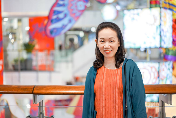 Frau in orangefarbenem Hemd und blauer Jacke steckt ihre Hand in die Tasche und lächelt mit Kamera in Einkaufszentrum.  - Foto, Bild