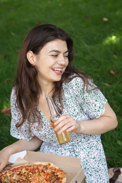 Happy young woman eating pizza and drinking a drink in a bottle at a picnic, copy space. - Fotoğraf, Görsel