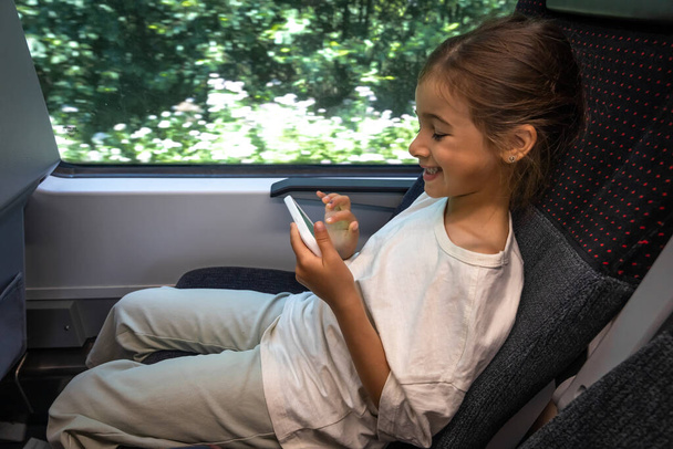 A little girl uses a smartphone while sitting in a train at the window. - Φωτογραφία, εικόνα