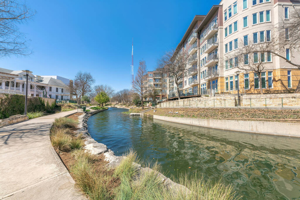 テキサス州サンアントニオのミッドライズの住宅街の真ん中にある小さな川。川の左側にはコンクリート製の歩道があり、右側の建物の奥には塔の景色が見えます。. - 写真・画像
