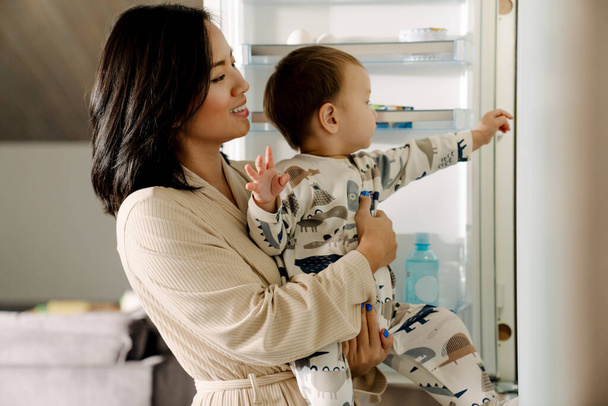 Happy mother holding her son while standing by open fridge at home - Фото, зображення