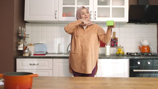 A hidzsábi nő valami zöldet tart a konyhában, megmutat egy terméket, mosolyog és egy vidám, képzeletbeli tárgyat mutat be. A kreatív 3D-s művészek bármilyen termékkel helyettesíthetik a zöld dobozt.. - Fotó, kép