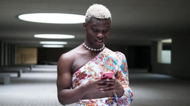 Стильна африканська чорна трансгендерна модель посміхається і пише смс на мобільному телефоні, стоячи в темному підземному гаражі. - Кадри, відео