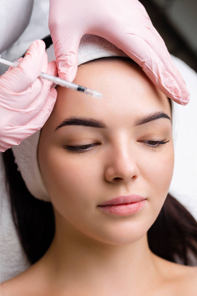 Close up of hands of young cosmetologist injecting botox in female face. She is standing and smiling. The woman is closed her eyes with relaxation. - Фото, изображение
