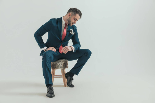 elegante hombre de negocios en traje comprobando su muñeca y sentado en silla de piel mientras posando sobre fondo gris en el estudio - Foto, imagen