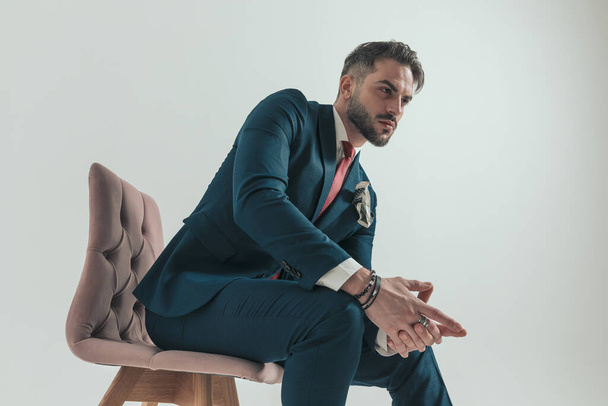 oldalnézet elegáns férfi öltönyben piros nyakkendővel és zsebkendővel ül rózsaszín bársony széken, és kéz a kézben, miközben keres oldalra - Fotó, kép