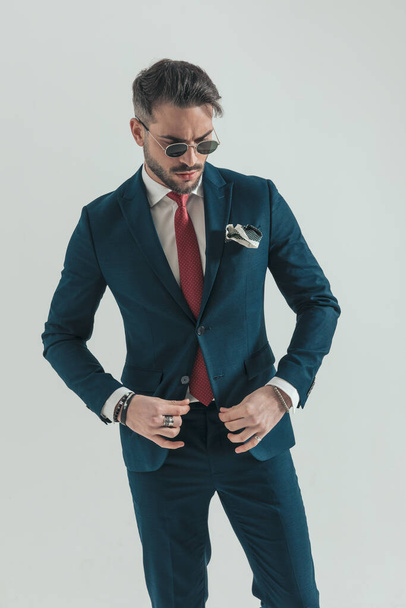 portrait of bearded elegant guy looking down and buttoning suit while posing on grey background in studio - Foto, Bild