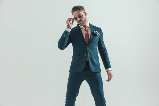 portrét cool módní muž v elegantním obleku pózovat sebevědomě při upevnění sluneční brýle před šedým pozadím ve studiu - Fotografie, Obrázek