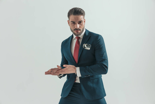 bel homme d'affaires portant un costume élégant avec cravate rouge et mouchoir, toucher et frotter les mains tout en posant devant fond gris - Photo, image