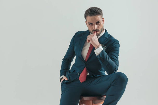 sinnlicher junger Mann im eleganten Anzug, der eine Hand in der Tasche hält und die Lippen auf verführerische Weise auf grauem Hintergrund im Studio berührt - Foto, Bild