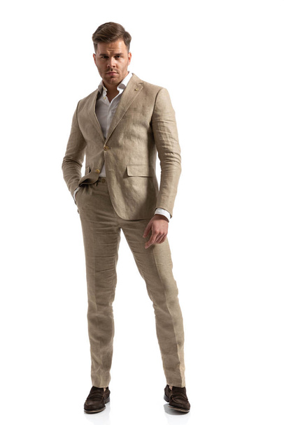 полное фото привлекательного мужчины в костюме в открытой рубашке с воротником, позирующего с рукой в кармане, стоя изолированно на белом фоне в студии - Фото, изображение