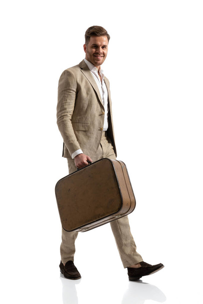 счастливый бизнесмен в костюме улыбается, держит багаж, берет несколько выходных и путешествует по миру, позирует на белом фоне в студии, все тело - Фото, изображение